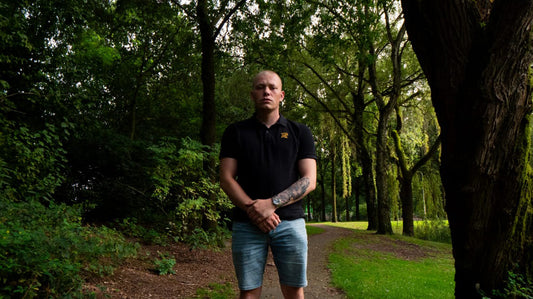 Rapper Nikkos1312 poseert met gekruiste armen in een park, dragend een zwarte Hellevated Polo, met gras en bomen op de achtergrond